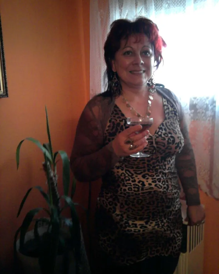 Femme aux cheveux roux portant un haut léopard et tenant un verre, debout près d'une plante.