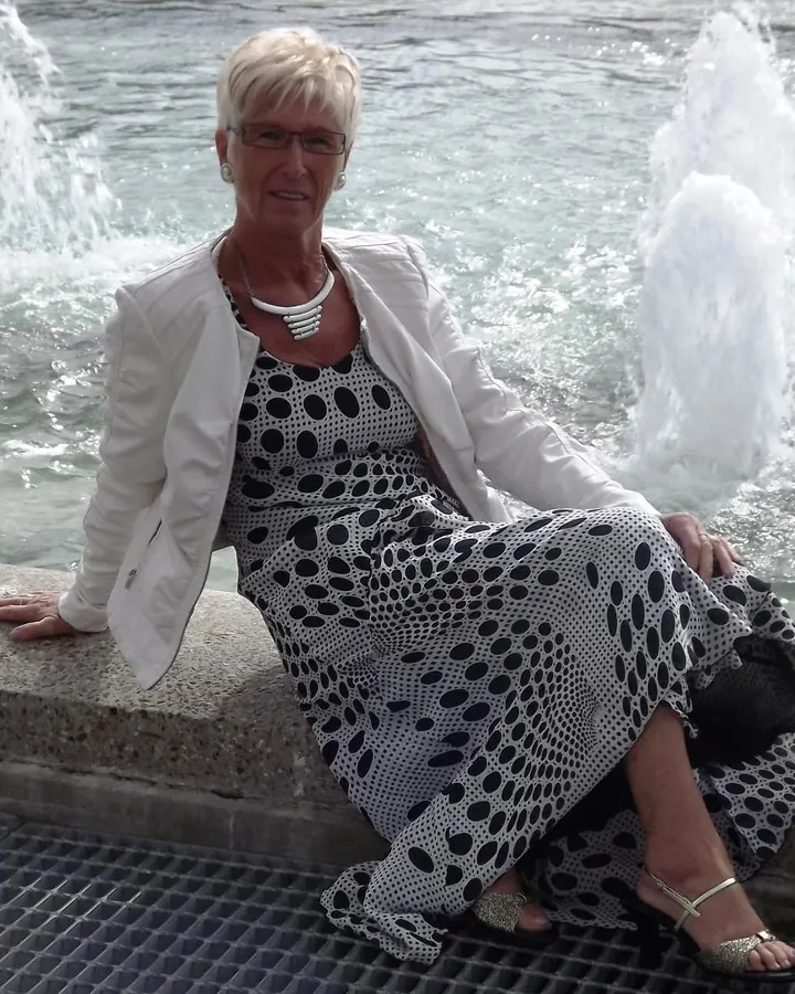 Femme aux cheveux blancs portant une veste blanche et une robe à pois noirs, assise près d'une fontaine.