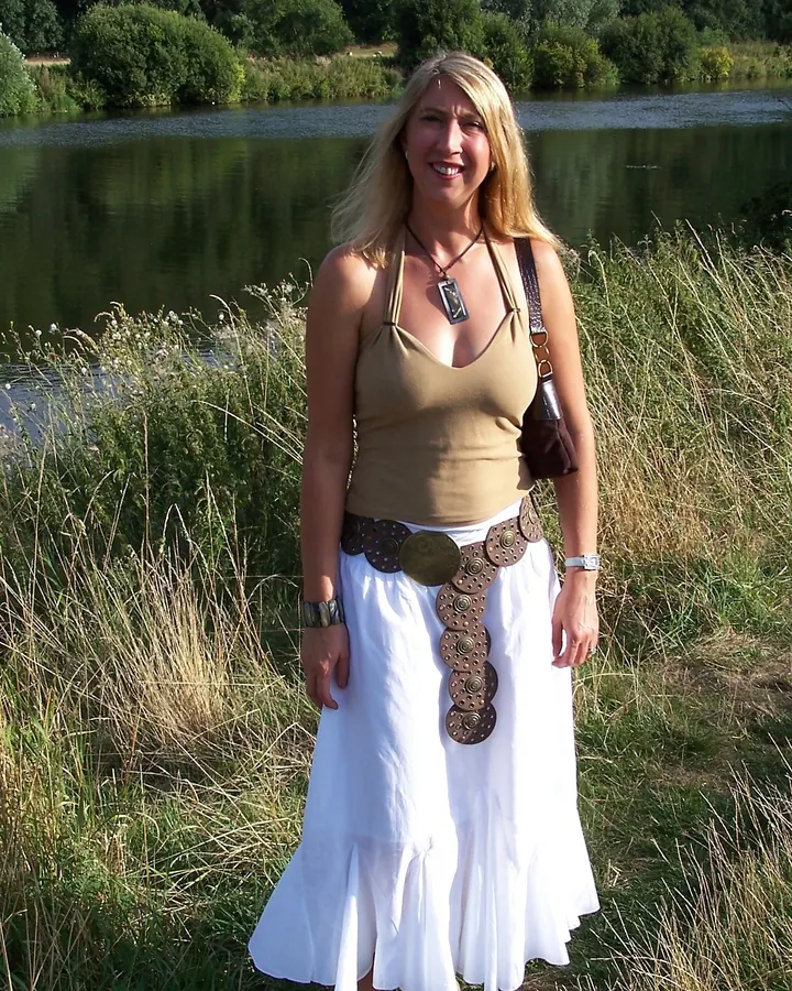 Femme blonde souriante portant un haut beige et une jupe blanche, debout près d’un lac.