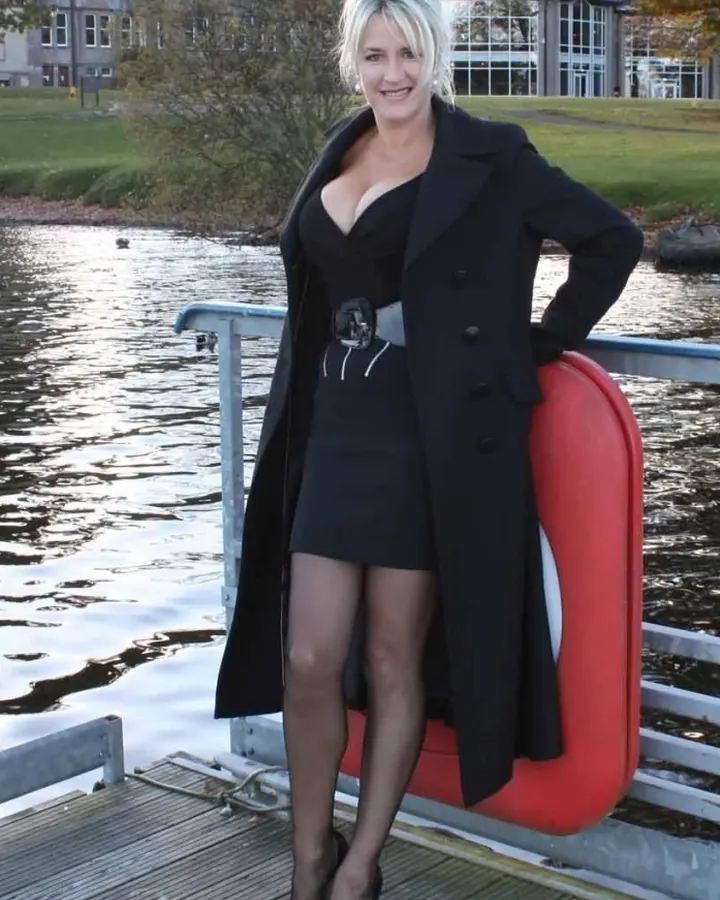 Femme blonde souriante, debout sur un pont, portant un manteau noir, une robe noire et des chaussures à talons.