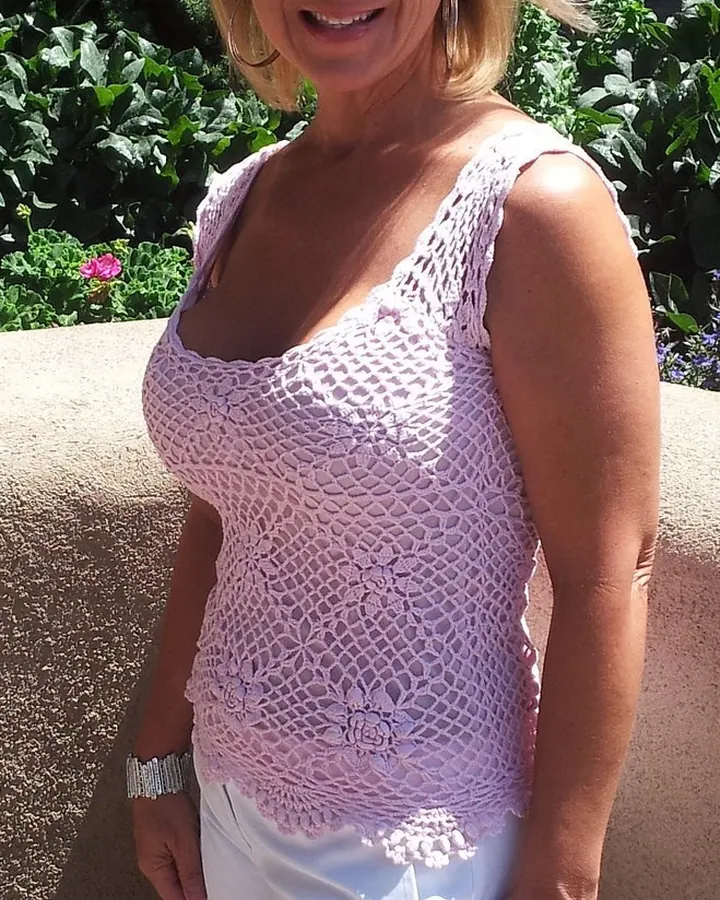 Femme blonde souriante, debout en extérieur, portant un haut rose crochet et un short blanc.
