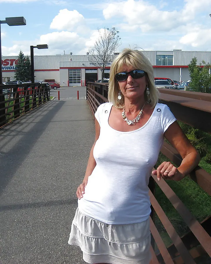 Femme blonde souriante, debout sur un pont, portant un haut blanc et une jupe blanche.